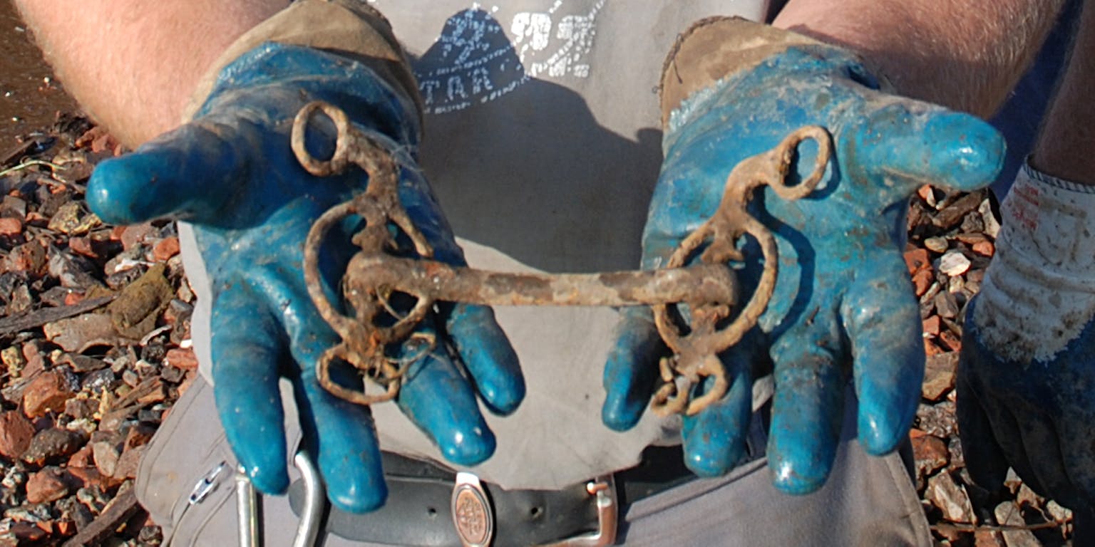 Een man met blauwe handschoenen toont een verroest paardenbit.