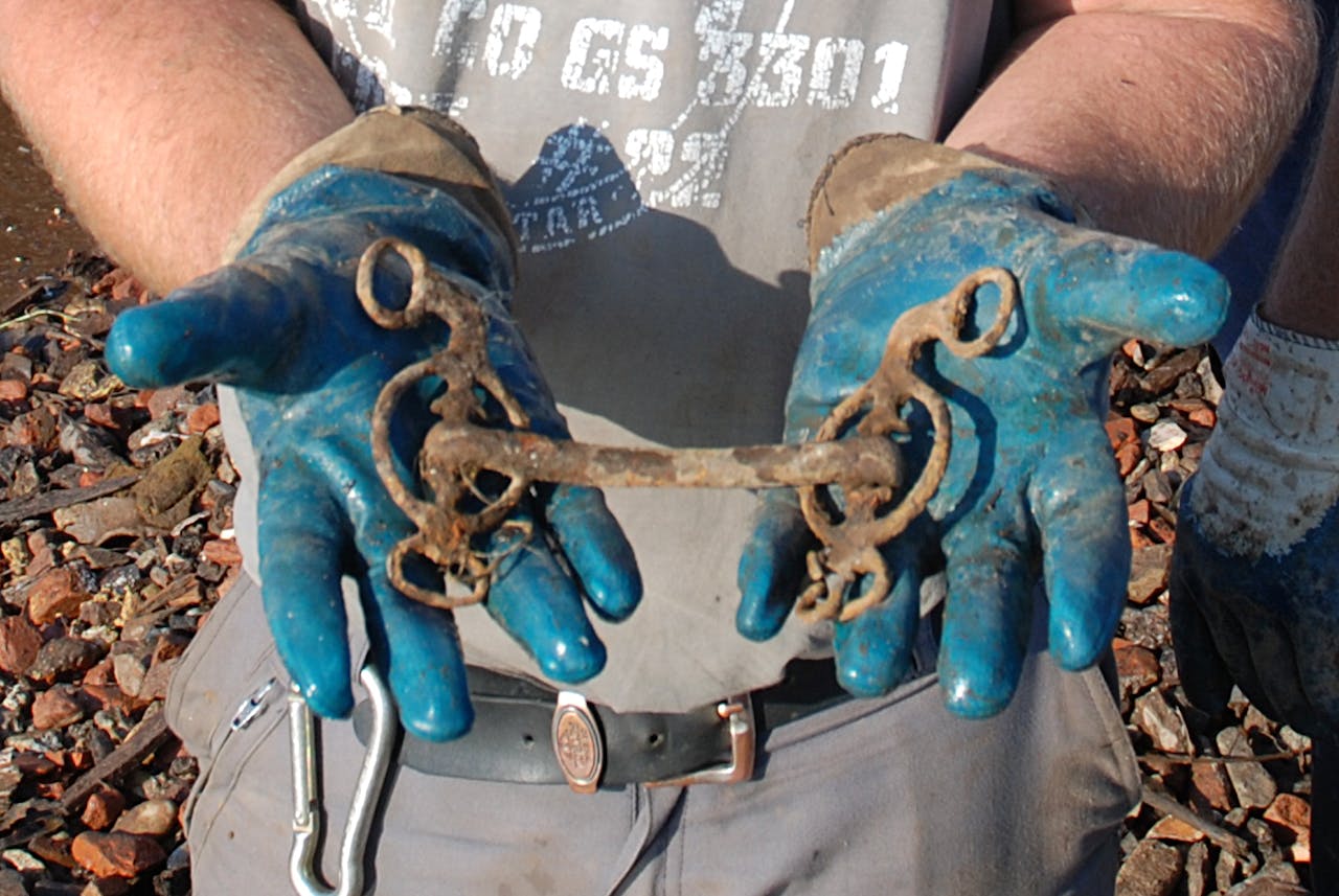 Een man met blauwe handschoenen toont een verroest paardenbit.