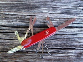 Een rood zakmes met uitgeklapte tools, waaronder een schaar, kurkentrekker, mes en opener, op een houten ondergrond.