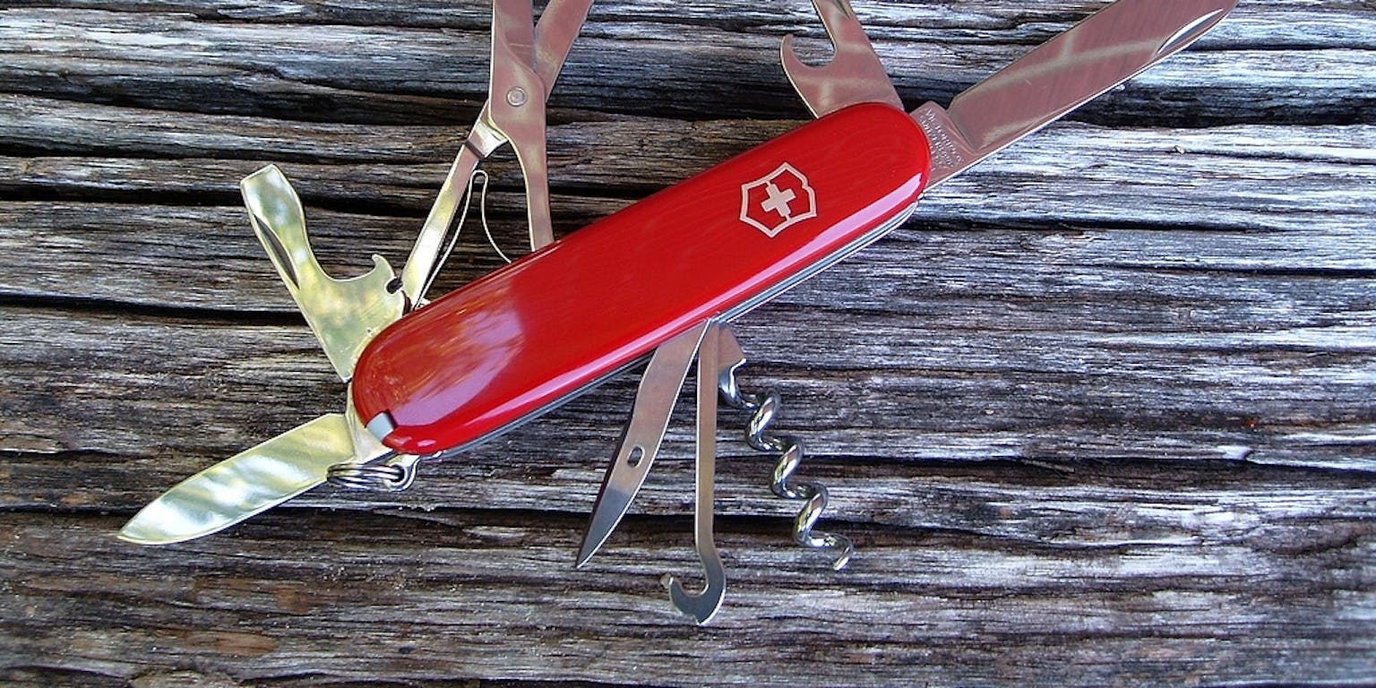 Een rood zakmes met uitgeklapte tools, waaronder een schaar, kurkentrekker, mes en opener, op een houten ondergrond.