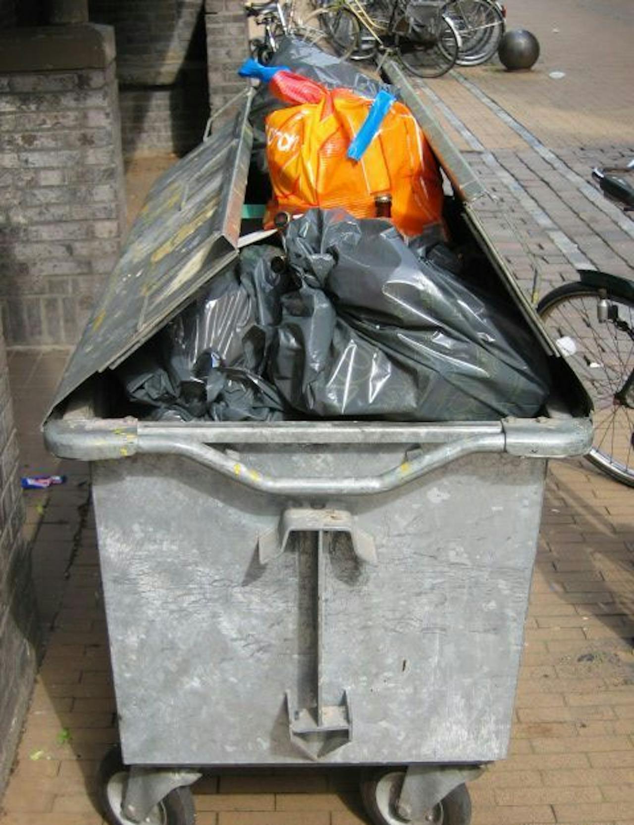 Een foto van een vuilniscontainer met allemaal grijze zakken en één oranje zak.