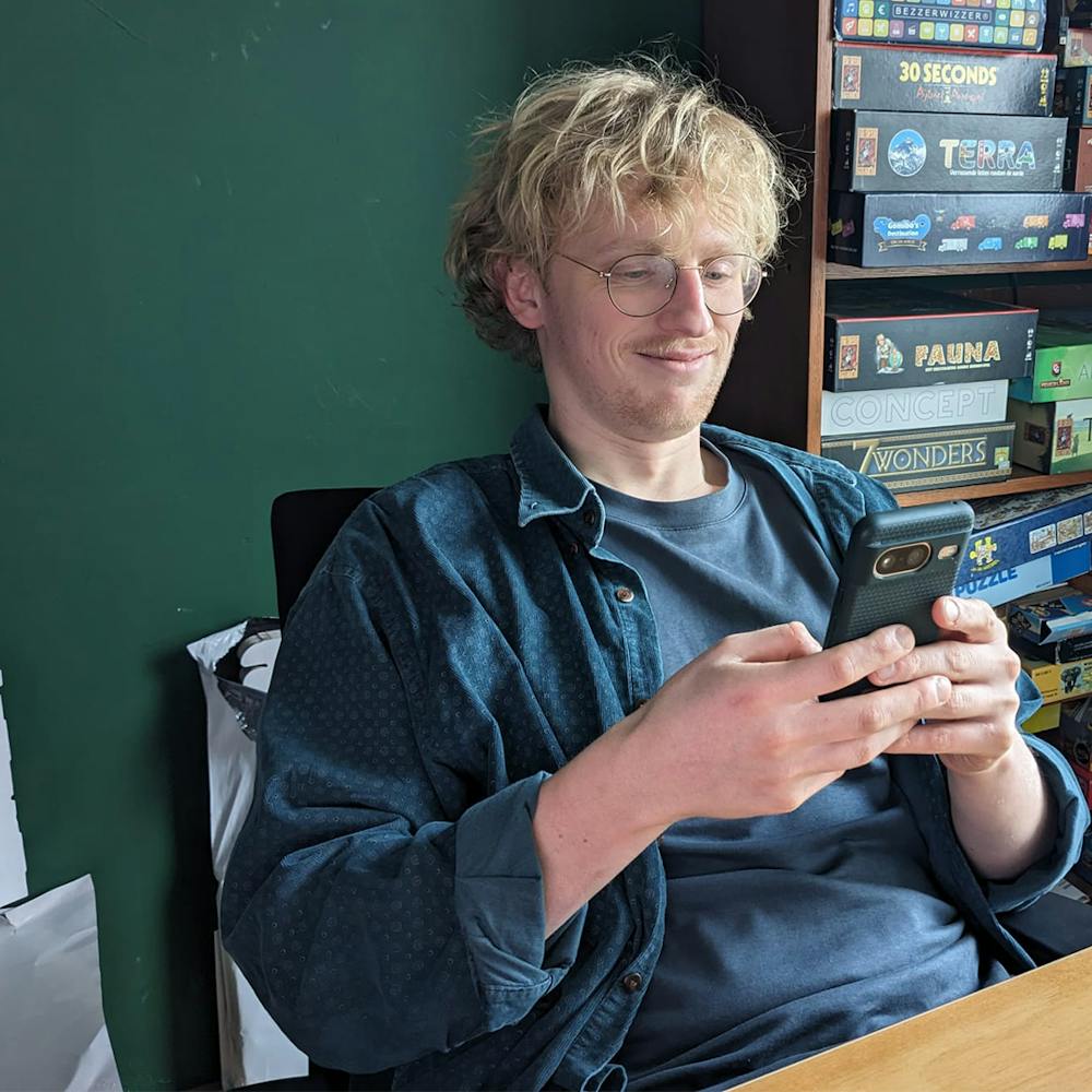 Kas Jansma bekijkt, zittend aan zijn bureau, glimlachend een meme op de smartphone in zijn handen.