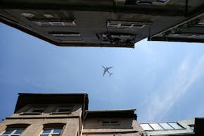 Tussen twee rijen gevels van gebouwen is vanaf de grond een vliegtuig te zien tegen een blauwe lucht.