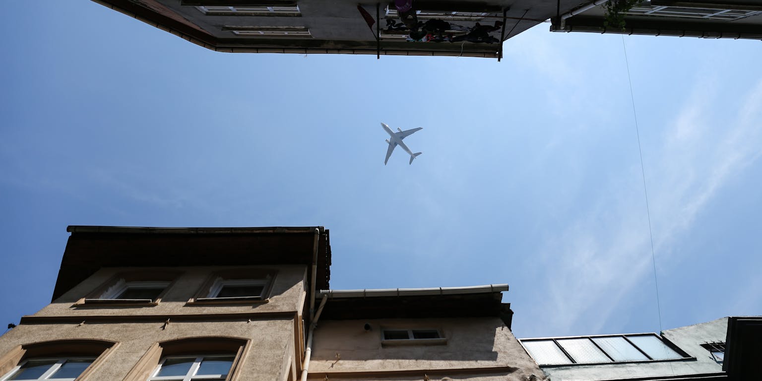 Tussen twee rijen gevels van gebouwen is vanaf de grond een vliegtuig te zien tegen een blauwe lucht.