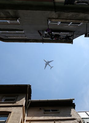 Tussen twee rijen gevels van gebouwen is vanaf de grond een vliegtuig te zien tegen een blauwe lucht.