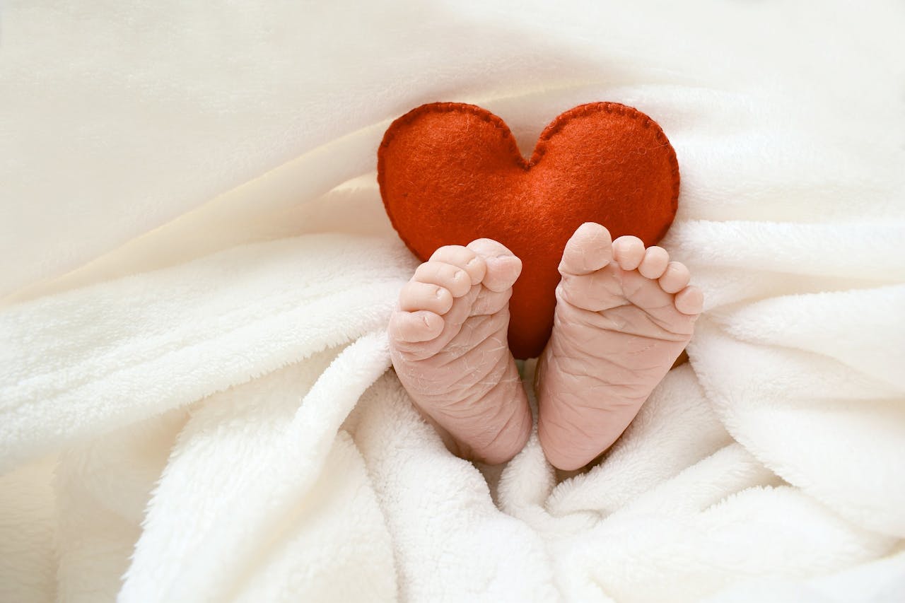 Een foto van een baby onder de dekens. Alleen de voetjes en een rood knuffelhart komen boven de dekens uit,