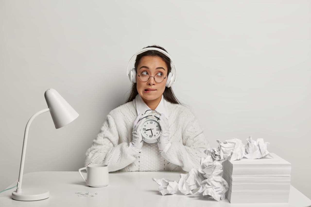 Jonge vrouw met witte kleding in witte omgeving houdt een witte wekker vast, ze kijkt een beetje zenuwachtig naar de wekker