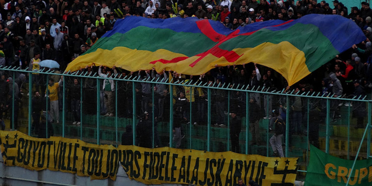 Een voetbaltribune in Algerije. Een grote groep mensen houdt een Berbervlag boven hun hoofd.