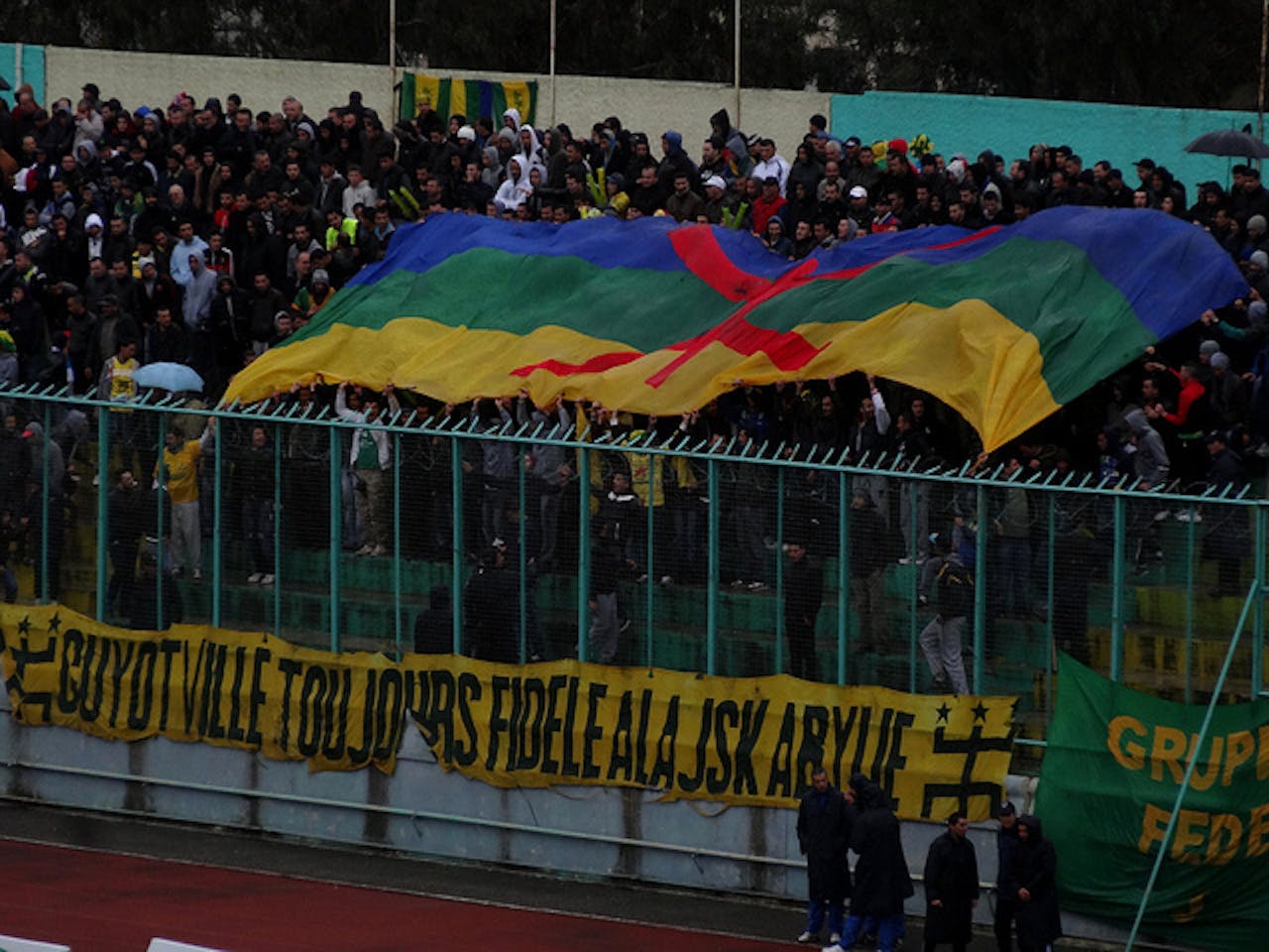 Een voetbaltribune in Algerije. Een grote groep mensen houdt een Berbervlag boven hun hoofd.
