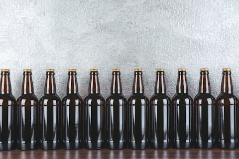 Nederland kende vroeger het standaardbierflesje, maar brouwerijen introduceren steeds vaker afwijkende flessen om zich te onderscheiden. Rij van bruine bierflesjes zonder etiket op een houten tafel tegen een grijs gemêleerde wand.
