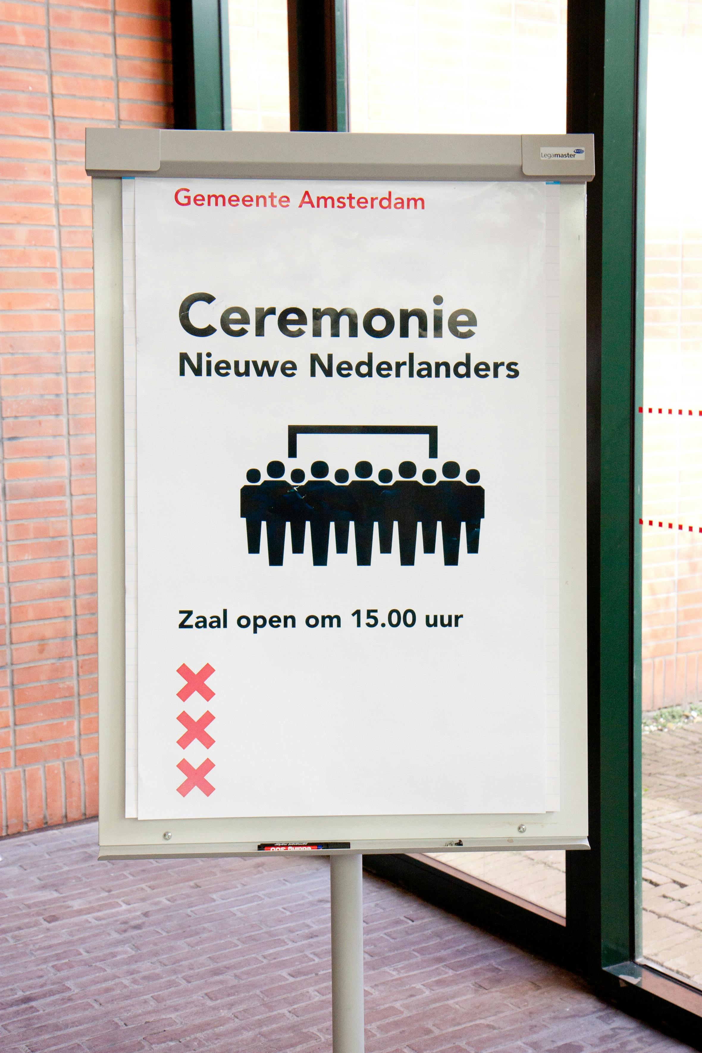 Een bord op een gebouw met de tekst: ceremonie nieuwe Nederlanders. Zaal open om 15:00 uur. De drie rode kruizen van Amsterdam zijn ook te zien. Ook zien we een groep met poppetjes.