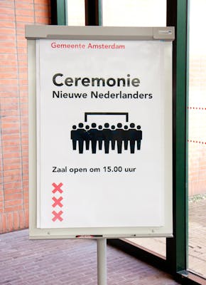 Een bord op een gebouw met de tekst: ceremonie nieuwe Nederlanders. Zaal open om 15:00 uur. De drie rode kruizen van Amsterdam zijn ook te zien. Ook zien we een groep met poppetjes.