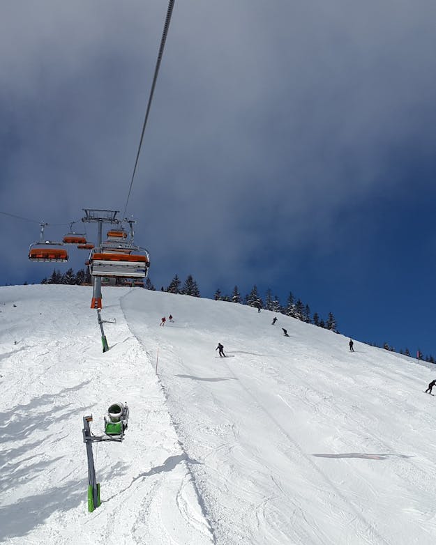 Een skipiste met een stoeltjeslift. De stoeltjes hebben een rode overkapping. Links en rechts staan besneeuwde dennenbomen. De lucht is blauw. Een skipiste met een stoeltjeslift. De stoeltjes hebben een rode overkapping. Links en rechts staan besneeuwde dennenbomen. De lucht is blauw.