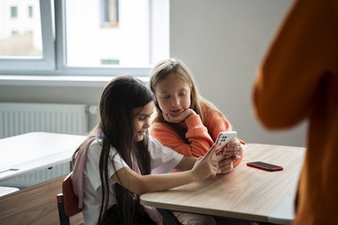 Twee meisjes zitten achter een tafeltje in een klaslokaal. Samen kijken ze op de telefoon van een van de meisjes. De telefoon van het andere meisje ligt op tafel.