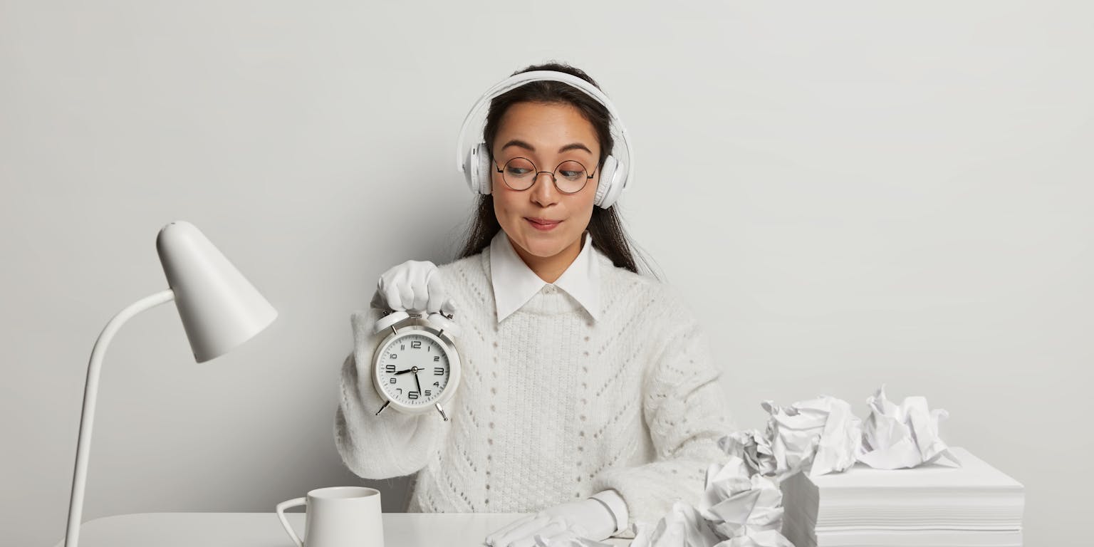 Jonge vrouw met witte kleding in witte omgeving van witte lamp, witte tafel en witte papieren en witte propjes papier houdt een witte wekker vast