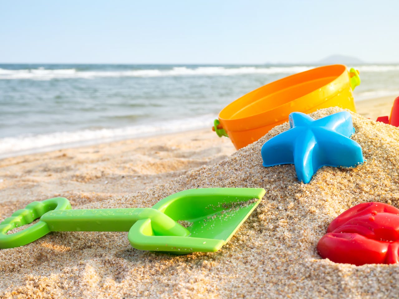 Een groen plastic schepje, blauwe zeestervorm, oranje emmer en rood vormpje liggen op het strand met op de achtergrond de zee.