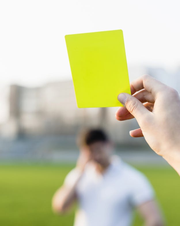 Een hand houdt een gele kaart vast. Op de achtergrond is wazig een voetbalveld te zien, met een speler die zichzelf voor z'n hoofd slaat. Een hand houdt een gele kaart vast. Op de achtergrond is wazig een voetbalveld te zien, met een speler die zichzelf voor z'n hoofd slaat.