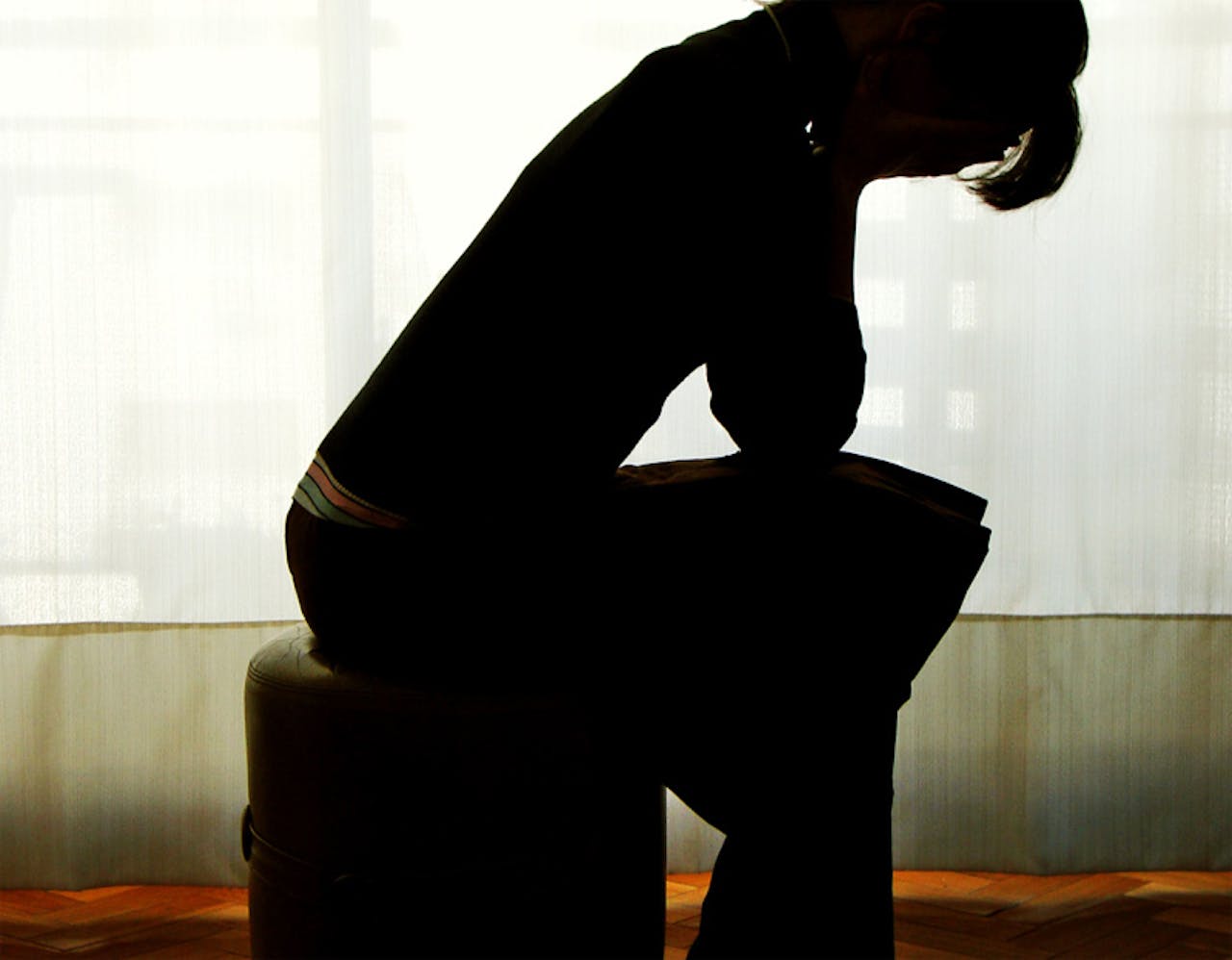 Een silhouet van een depresief persoon. Zit terneergeslagen bij een gordijn op een poef.