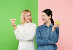 twee jonge vrouwen met een groene en roze achtergrond moeten kiezen tussen een gezonde appel eten, of een ongezond cakeje