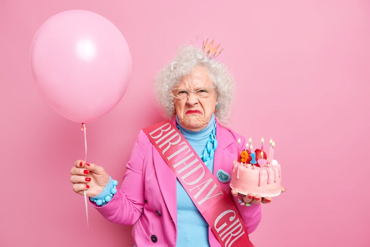 oude jarige vrouw met verjaardagstaart en roze ballon en sjerp met 'birthday girl' erop kijkt heel erg boos