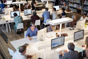 Verschillende mensen werkend achter een computer aan lange witte tafels in een bibliotheek.