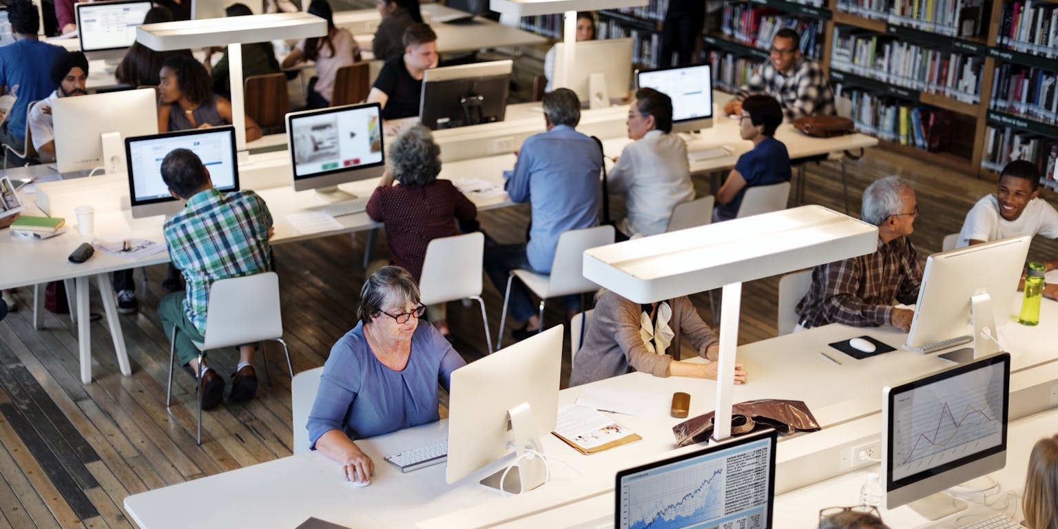 Verschillende mensen werkend achter een computer aan lange witte tafels in een bibliotheek.