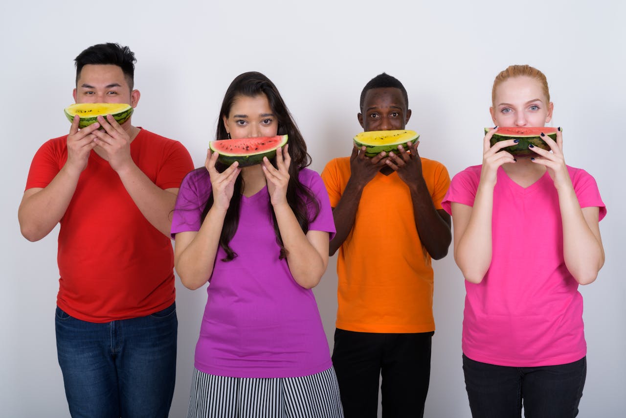 groep van vier jonge mensen van verschillende etniciteiten in kleurige t-shirts eten watermeloen en ananasschijven die ze voor hun mond houden