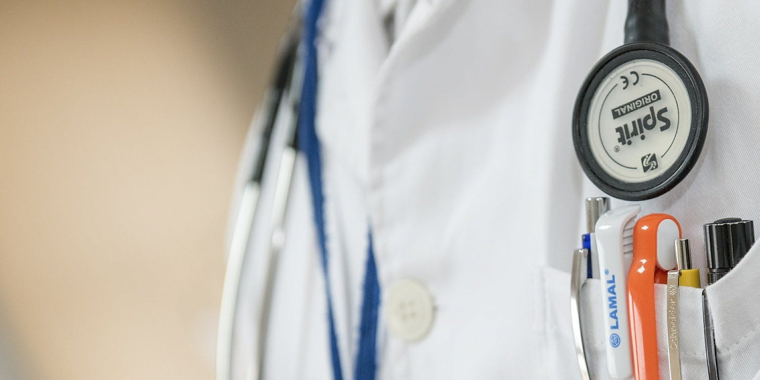 Een close-up van een doktersjas. Er is een stethoscoop te zien en een borstzakje vol met pennen.