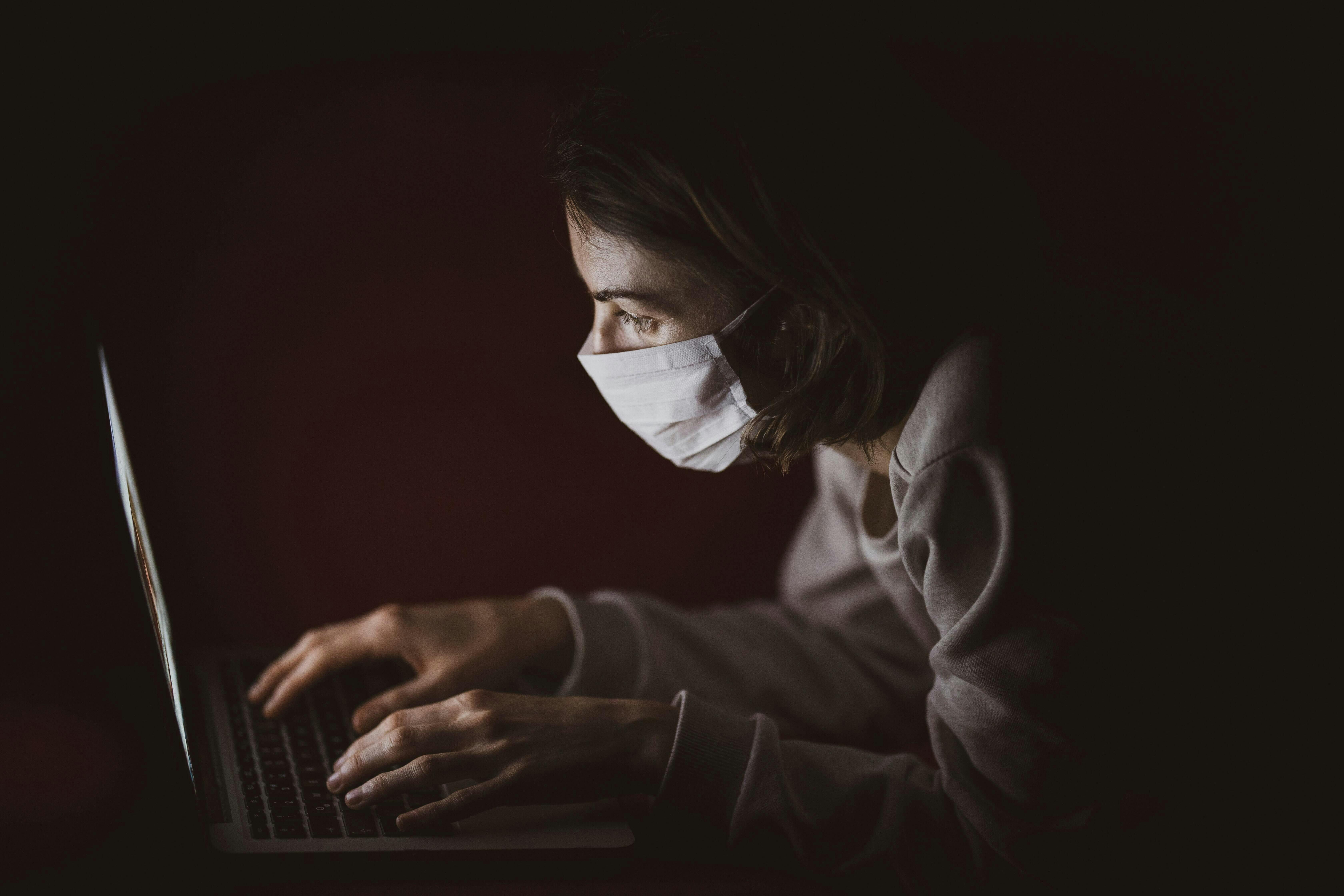 Een vrouw die een medisch masker draagt, gebruikt een laptop in het donker.