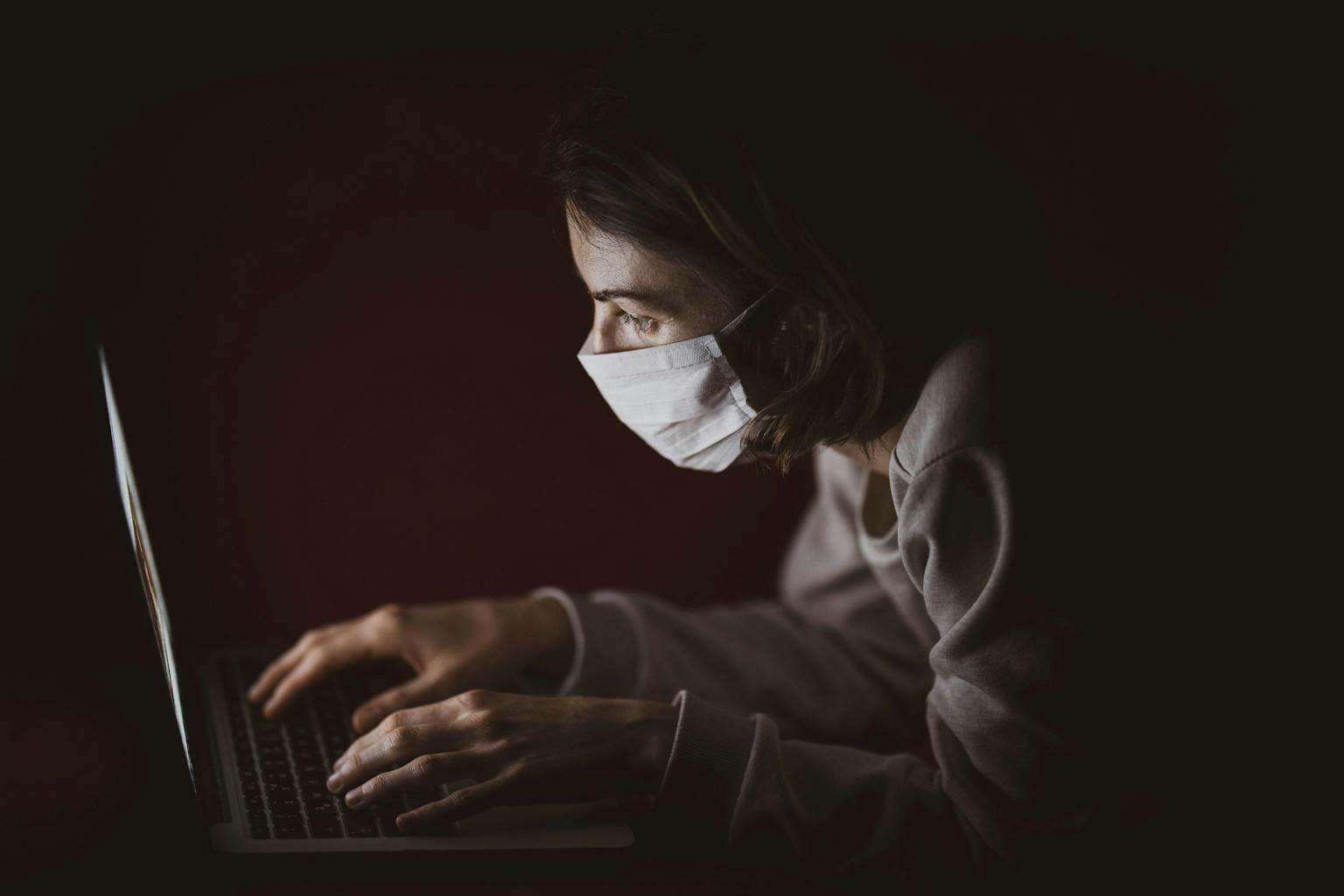 Een vrouw die een medisch masker draagt, gebruikt een laptop in het donker.