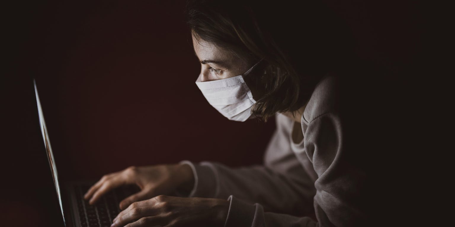 Een vrouw die een medisch masker draagt, gebruikt een laptop in het donker.