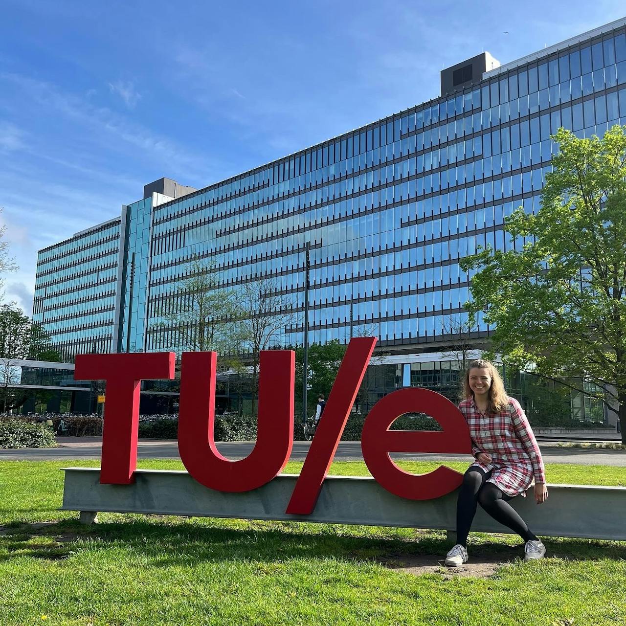 Femke Witmans op bezoek bij de TU Eindhoven, zittend vlakbij de rode letters met TUE voor het universiteitsgebouw