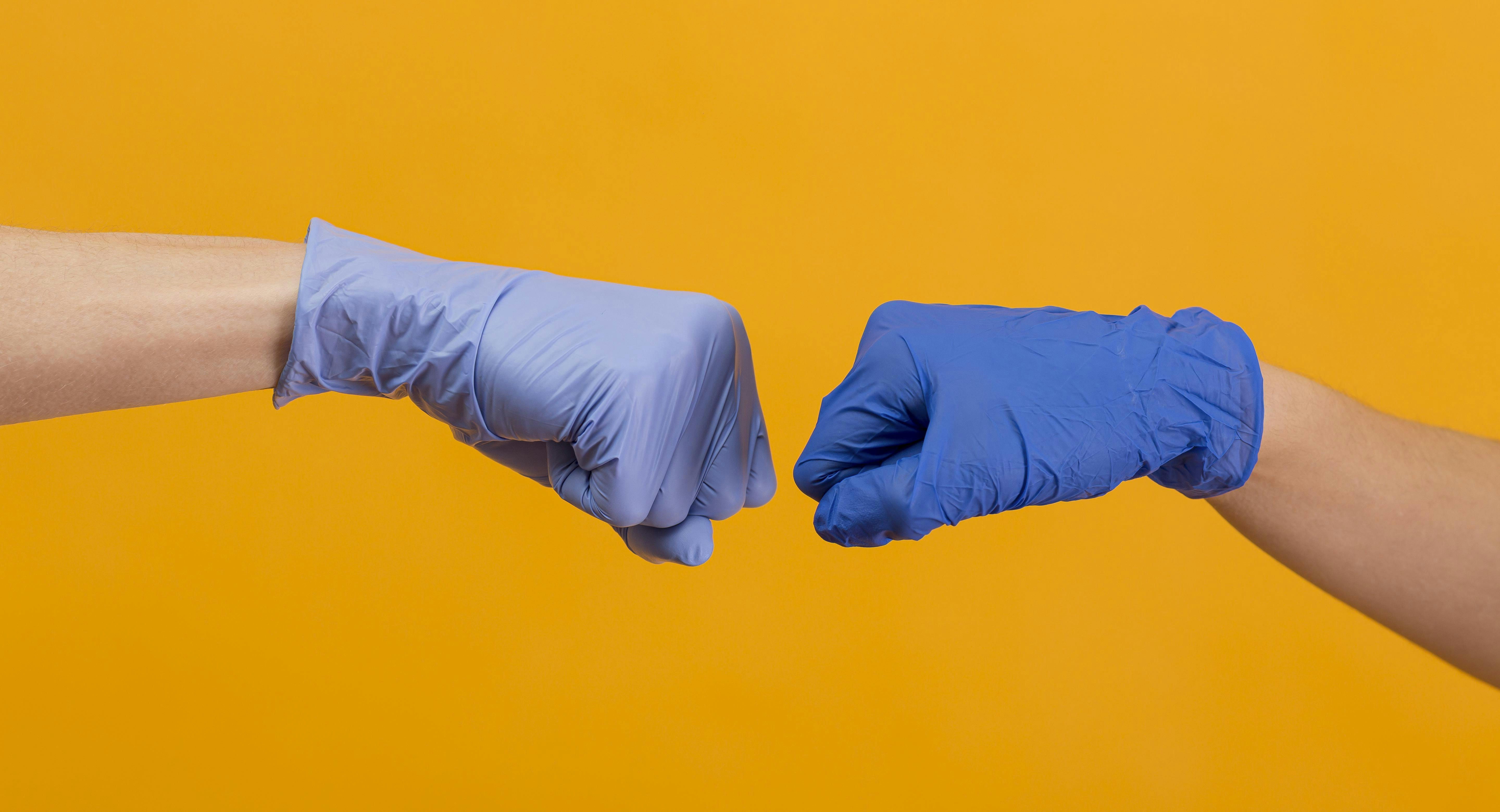 twee mensen geven elkaar een boks met blauwe plastic labhandschoenen aan op een gele achtergrond