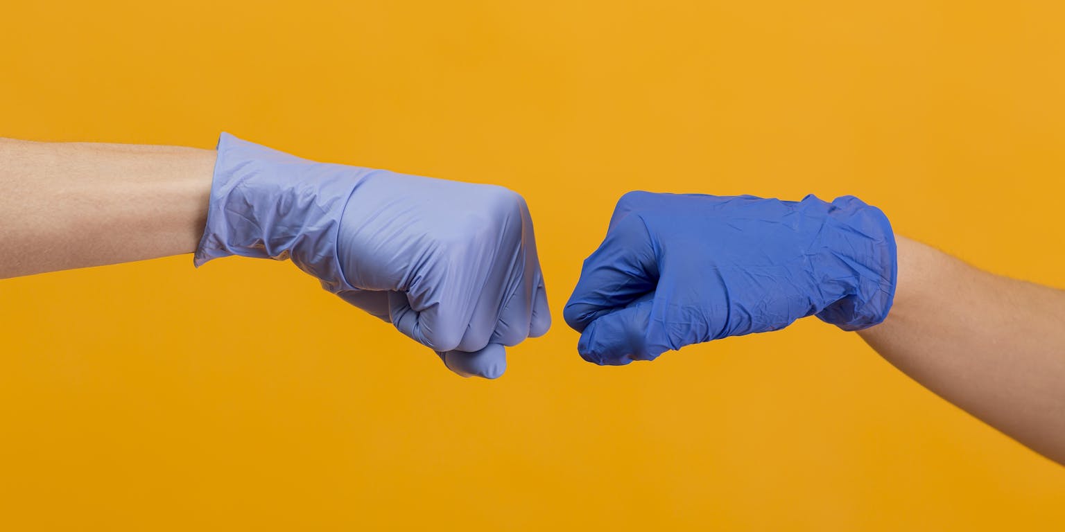 twee mensen geven elkaar een boks met blauwe plastic labhandschoenen aan op een gele achtergrond