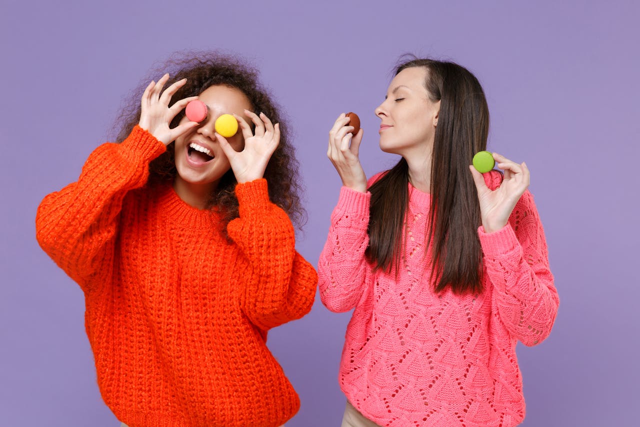 Twee tieners in rode en roze trui met paarse achtergrond spelen met macarons. Een tiener houdt de macarons als een bril voor haar ogen, de ander ruikt eraan