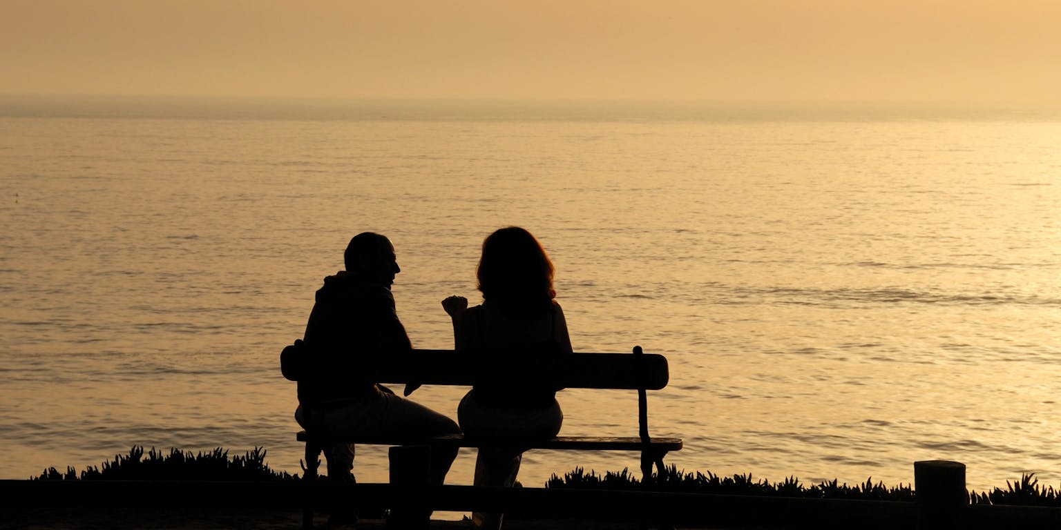 De silhouetten van twee mensen die bij zonsondergang een gesprek op een bankje voeren.