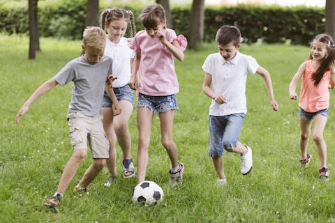 Kinderen zouden minimaal twee uur per dag buiten moeten spelen. Drie meisjes en twee jongens rennen achter een voetbal aan in een park.