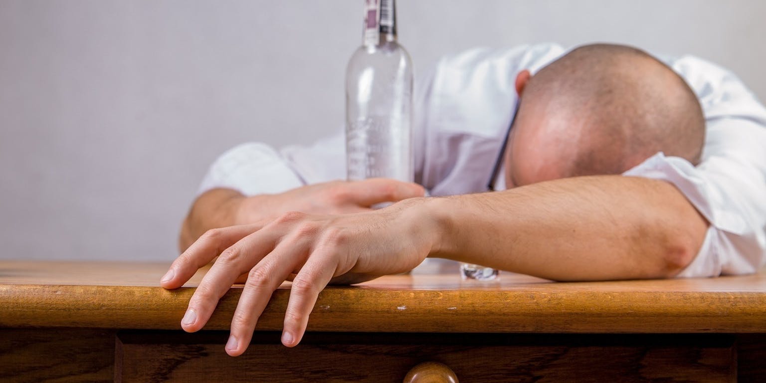 Een man slaapt op tafel met een fles alcohol in zijn hand.