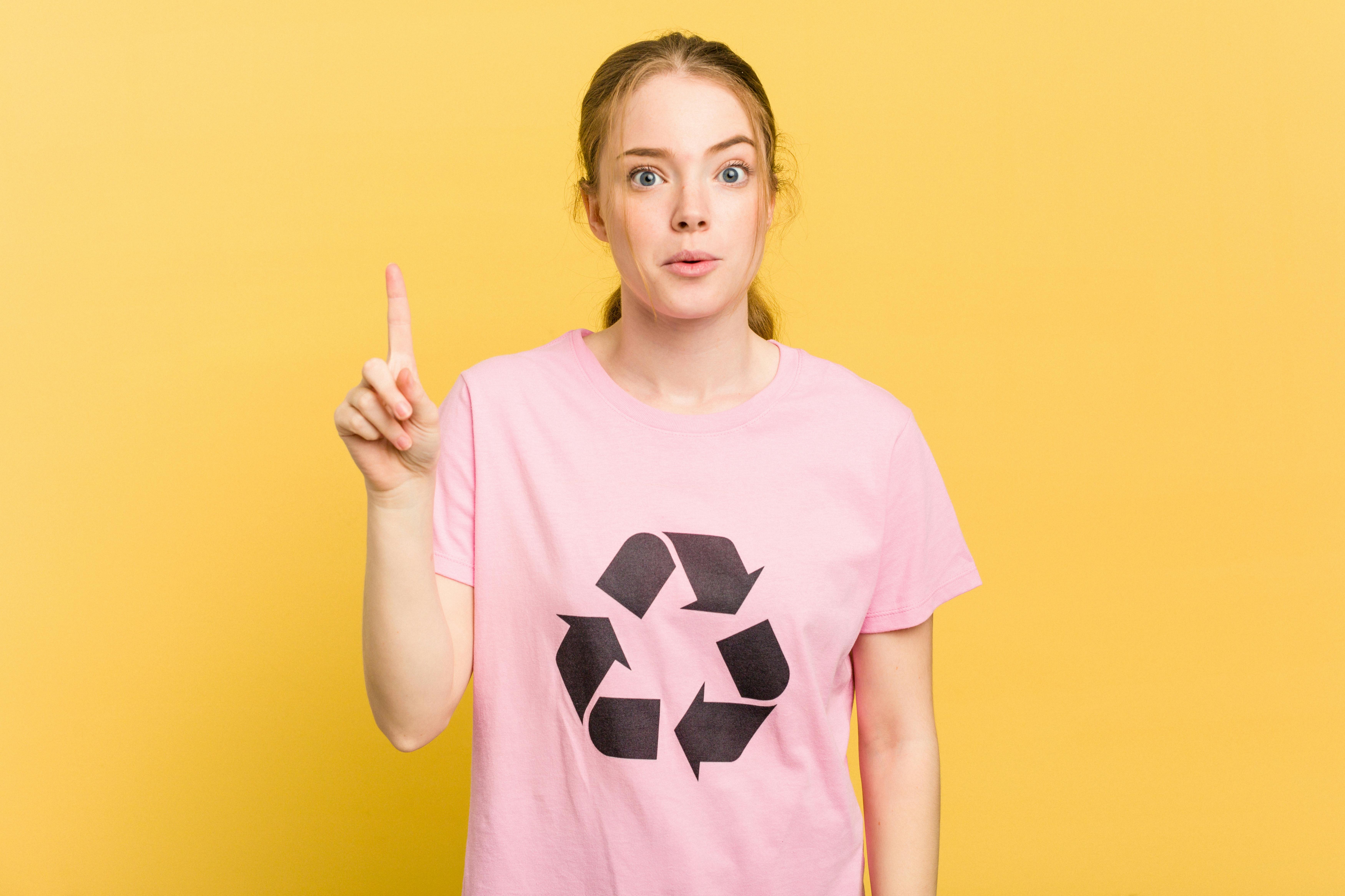 Jonge vrouw met roze shirt aan met recycleteken kijkt vragend en steekt haar vinger omhoog