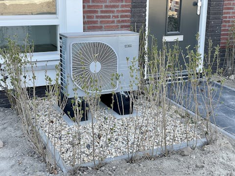Een buitenunit van een warmtepomp. In de voortuin van een huis staat een airconditioning unit.
