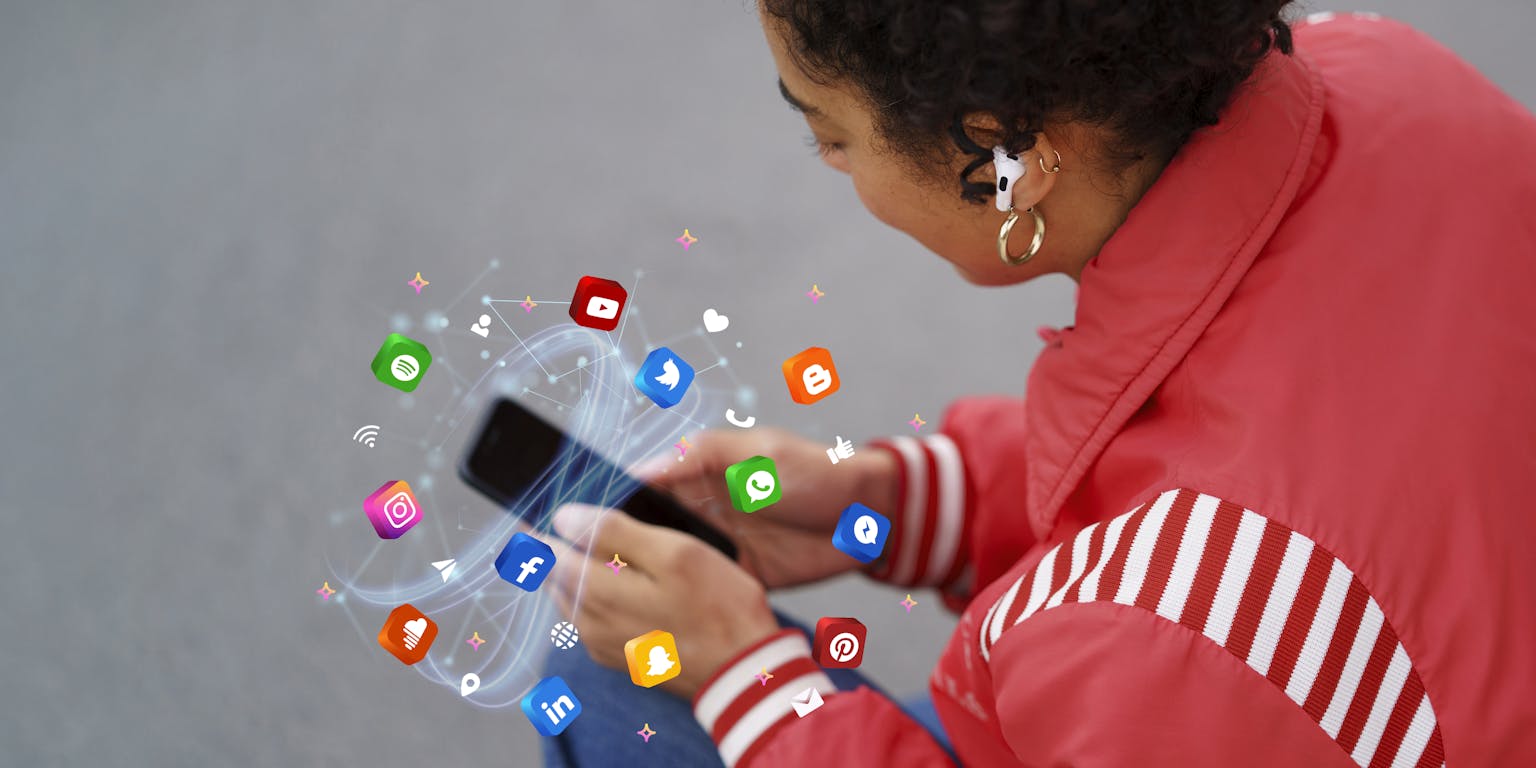 Een jongedame in een rode jas kijkt naar haar telefoon met oordopjes in. De camera kijkt over haar schouder. Rond de telefoon zweven icoontjes van verschillende apps.