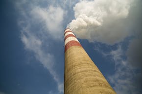 Betonnen schoorsteen met bovenaan rode en witte strepen gefotografeerd vanaf beneden tegen een blauwe lucht. uit de schoorsteen komt een witte rookwolk.