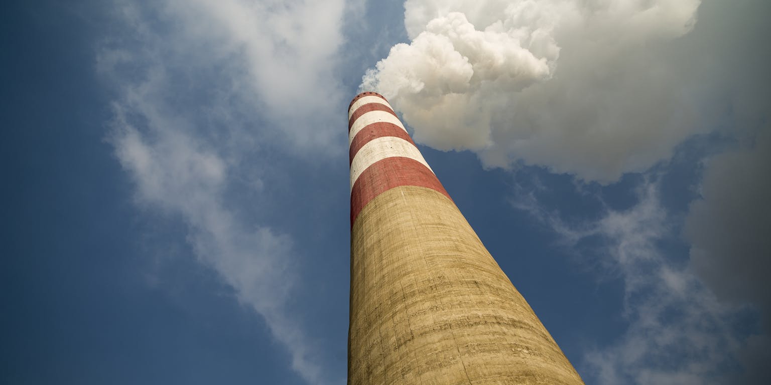 Betonnen schoorsteen met bovenaan rode en witte strepen gefotografeerd vanaf beneden tegen een blauwe lucht. uit de schoorsteen komt een witte rookwolk.