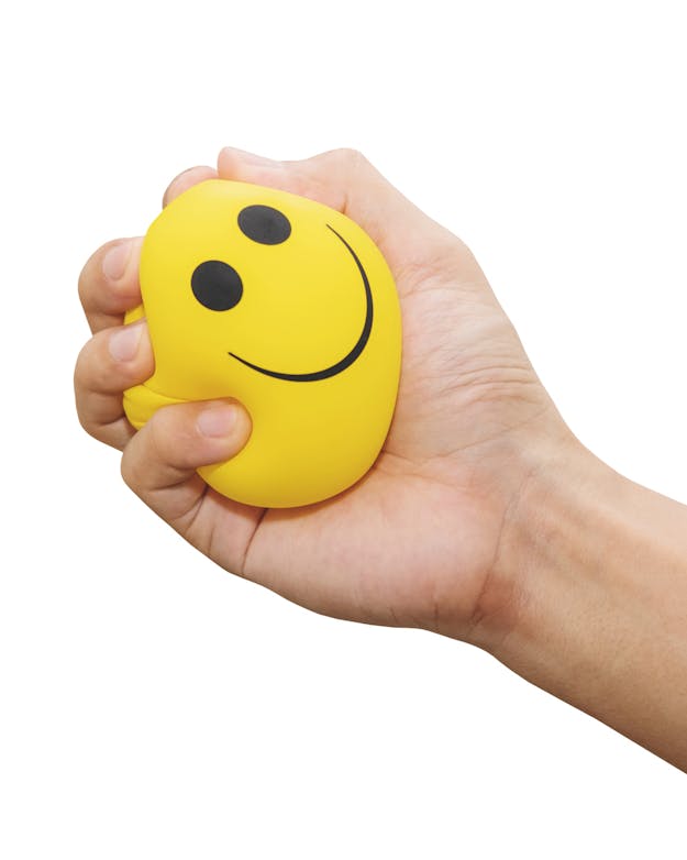 Hand die knijpt in een gele stressbal met een smiley erop. Hand die knijpt in een gele stressbal met een smiley erop.