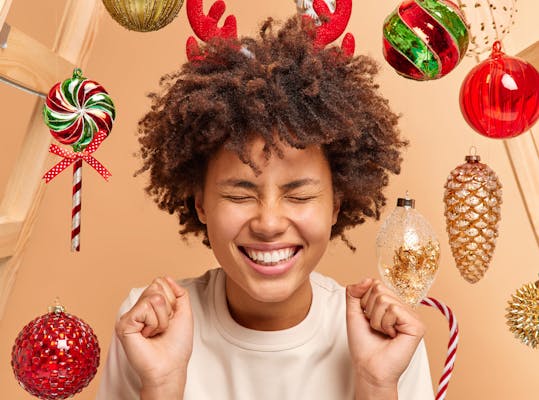 Jonge vrouw kijkt blij en tegelijkertijd wat gespannen of ze wel cadeautjes krijgt met kerst, om haar heen hangen rode en groene kerstballen en versieringen