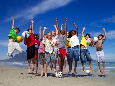 Een groep kinderen springt met voetballen op het strand.