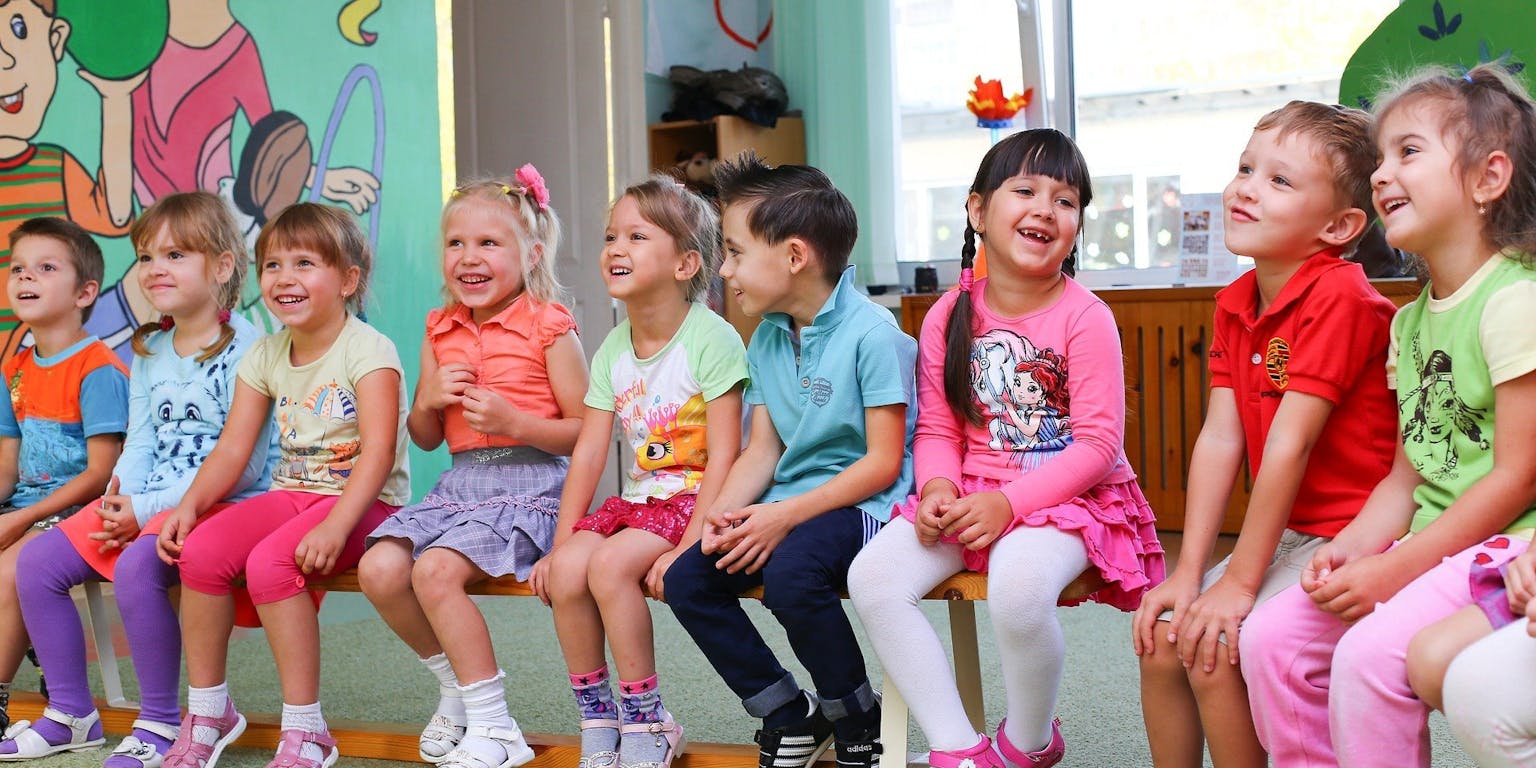 Een groep blije kinderen zit op een bankje in een klaslokaal.