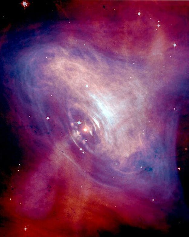 Het centrum van de Krabnevel. Hier bevindt zich een neutronster die eens in de 0,033 seconden ronddraait. De ster en de nevel ontstonden na een supernova-explosie in het jaar 1054. Een paars en rood gekleurde nevel tegen een zwarte sterrenhemel.