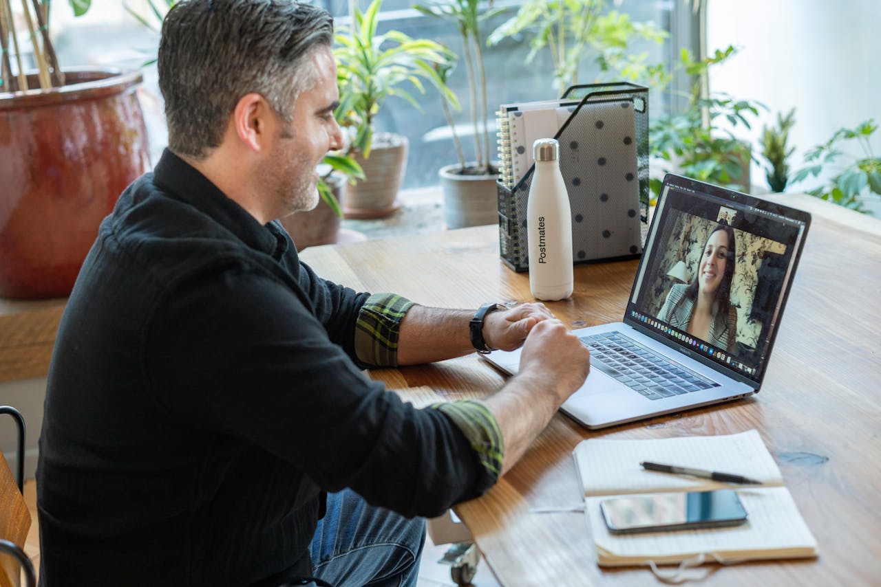 Een man zit aan een tafel met een laptop en zit in een videocall.
