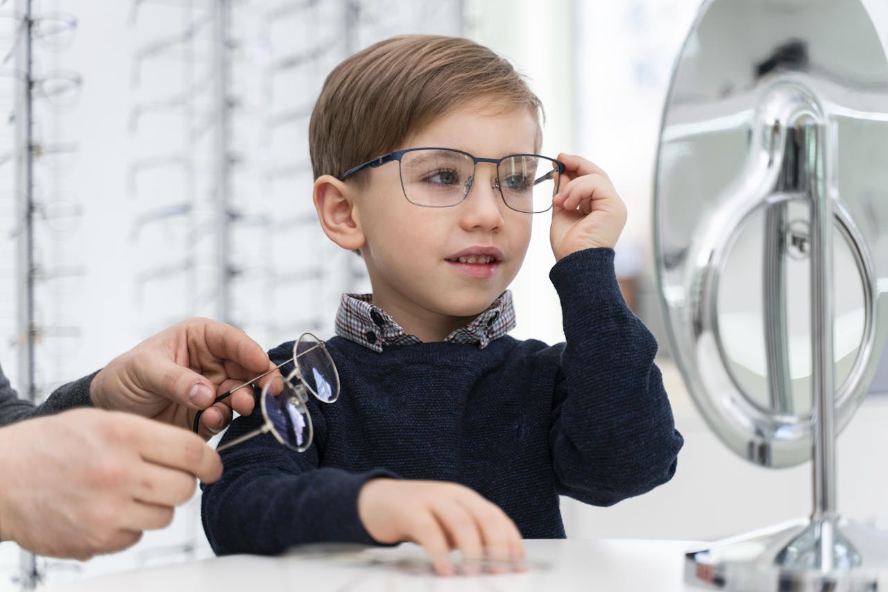 Kinderen in de groei hebben soms elk jaar een nieuwe bril nodig. Een jongetje past in een brillenwinkel een montuur en kijkt daarbij in een spiegel. Twee handen geven hem een tweede montuur aan.